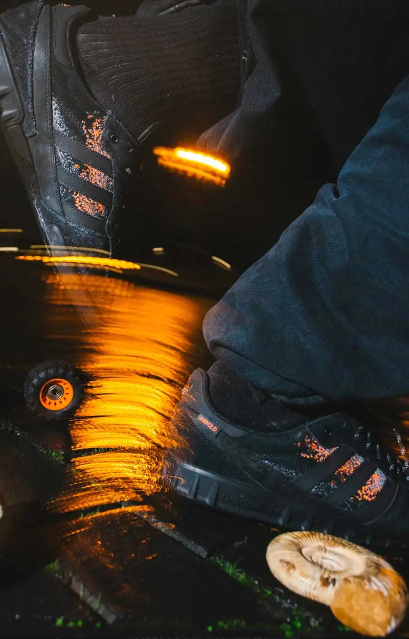 Schwarzer adidas Skateboarding Schuh mit orangefarbenen Details auf nassem Untergrund bei Nacht, aufgenommen in Bewegung